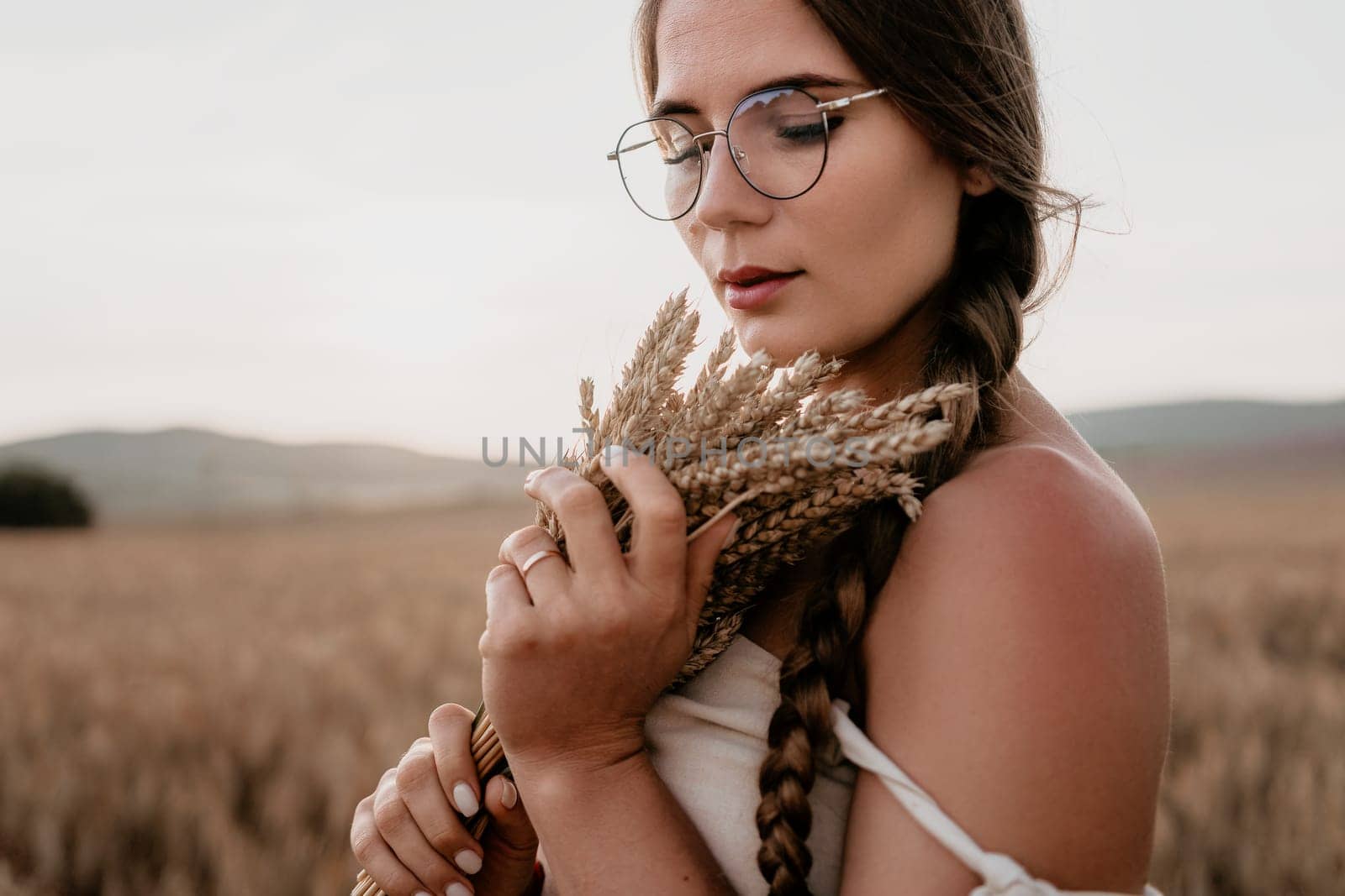 A young woman with long brown hair in braids holds wheat in a field, her eyes closed as she enjoys the moment.