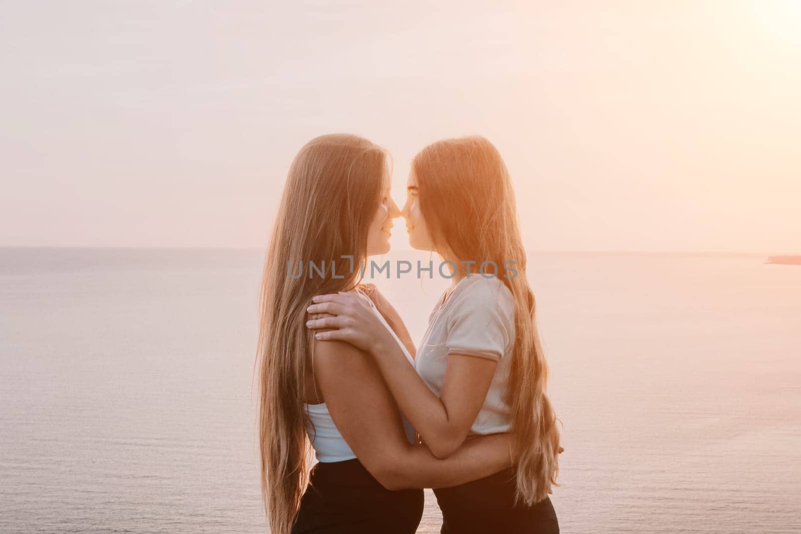 Two young women with long brown hair embrace in front of a tranquil ocean and sunset sky.