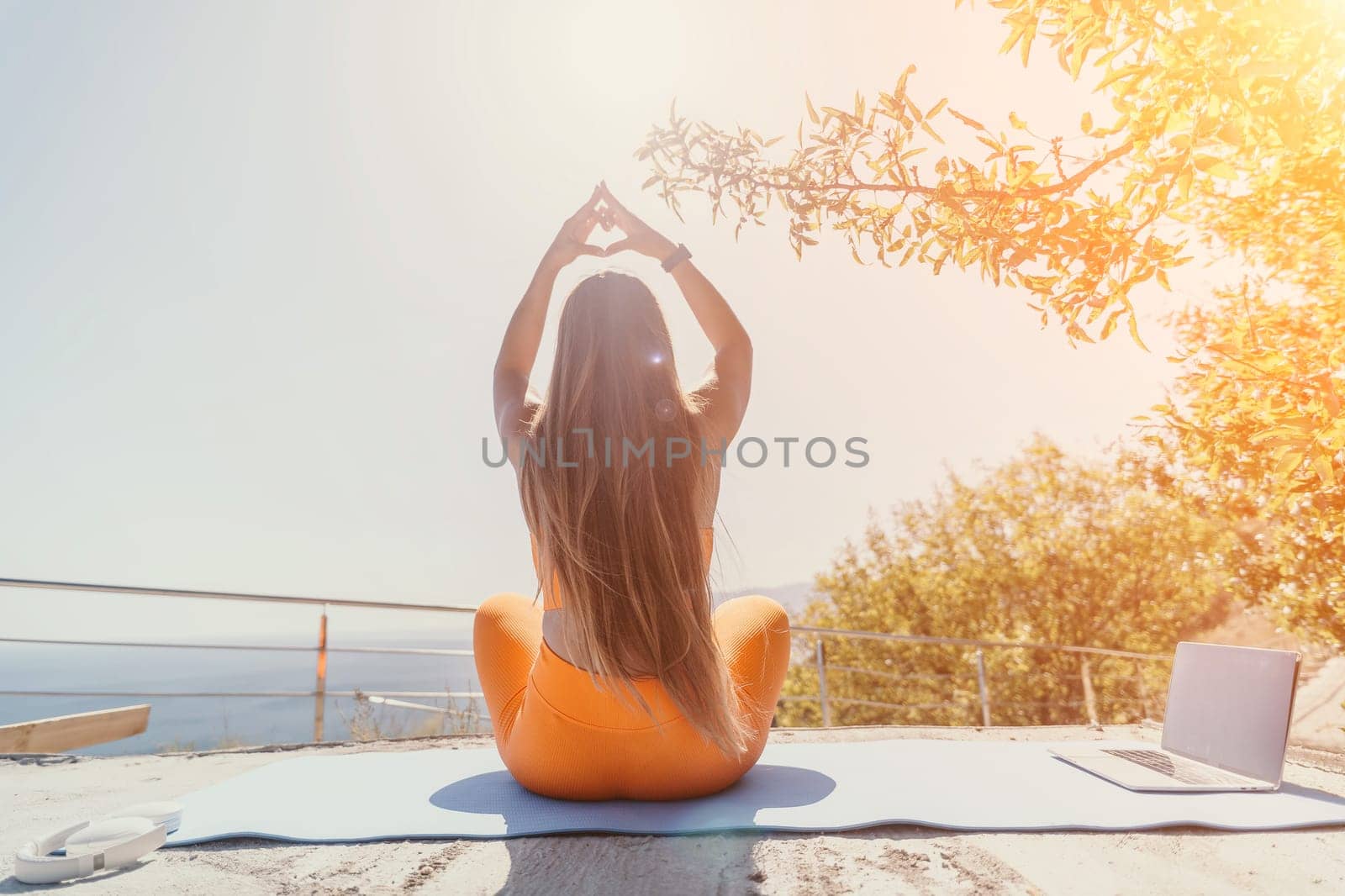 A young woman in orange sportswear sits in a yoga pose with her hands forming a heart shape, practicing outdoors with a laptop nearby.