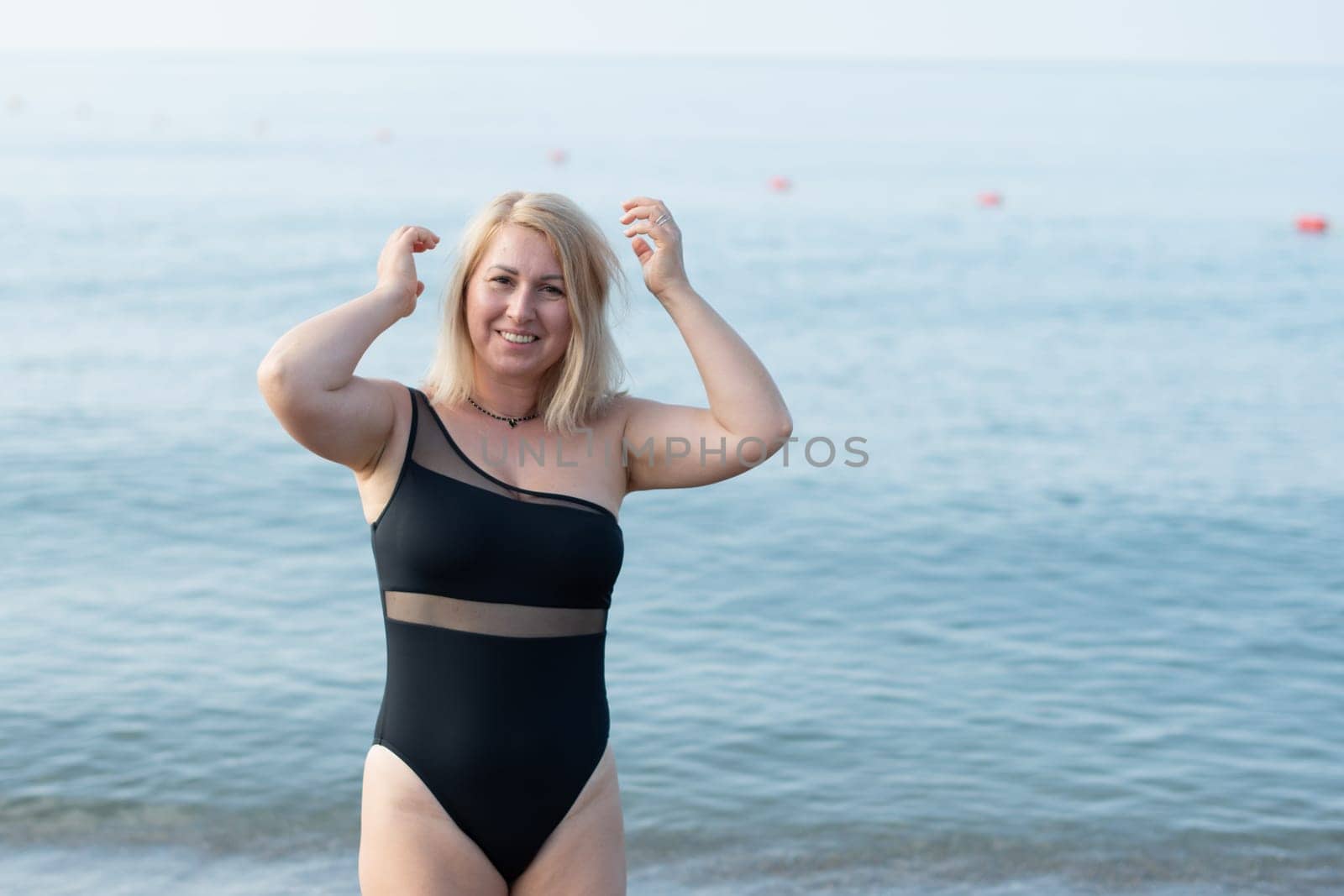 happy young woman in a fashionable black swimsuit stands on a sandy beach by the sea on a beautiful sunny day, straightening her wind-tumbled hair