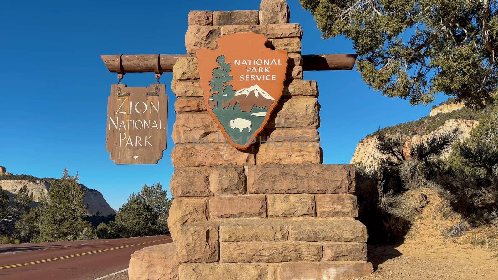East Zion, Utah, USA – 02.28.2024: Close up of the Zion National Park east entrance sign. It is a beautiful day with blue sky. The sign is made of stone and wood. It is located on the side of a road.
