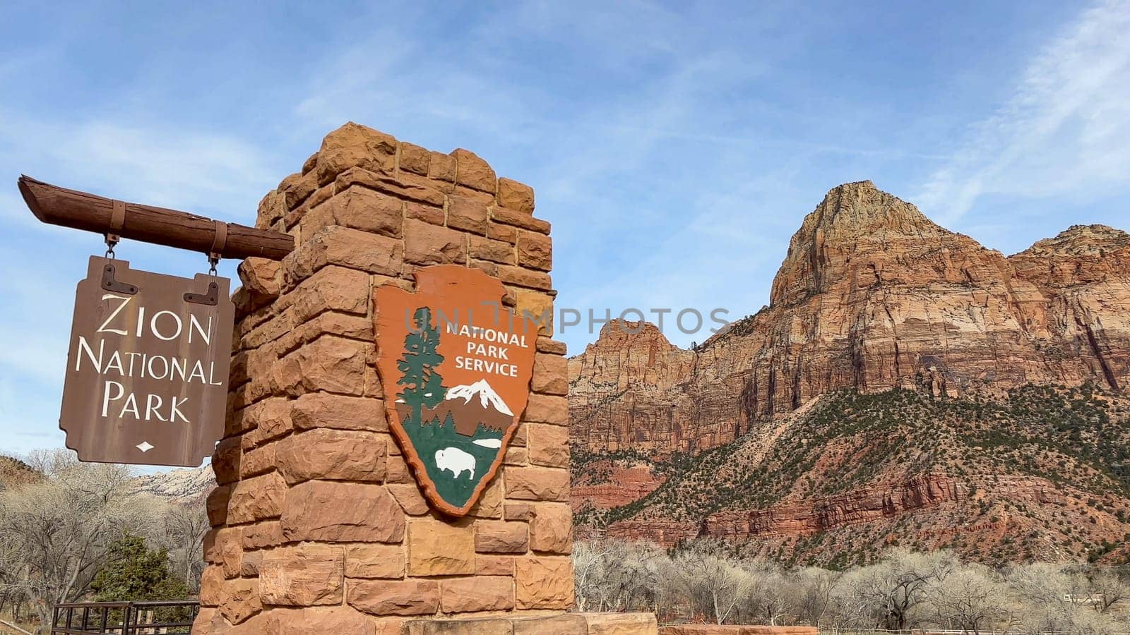 Springdale, Utah, USA - 02.27.2024: The main entrance sign for Zion National Park in Utah. The red rock sign is set against a beautiful blue sky, with the majestic Watchman mountain in the background.