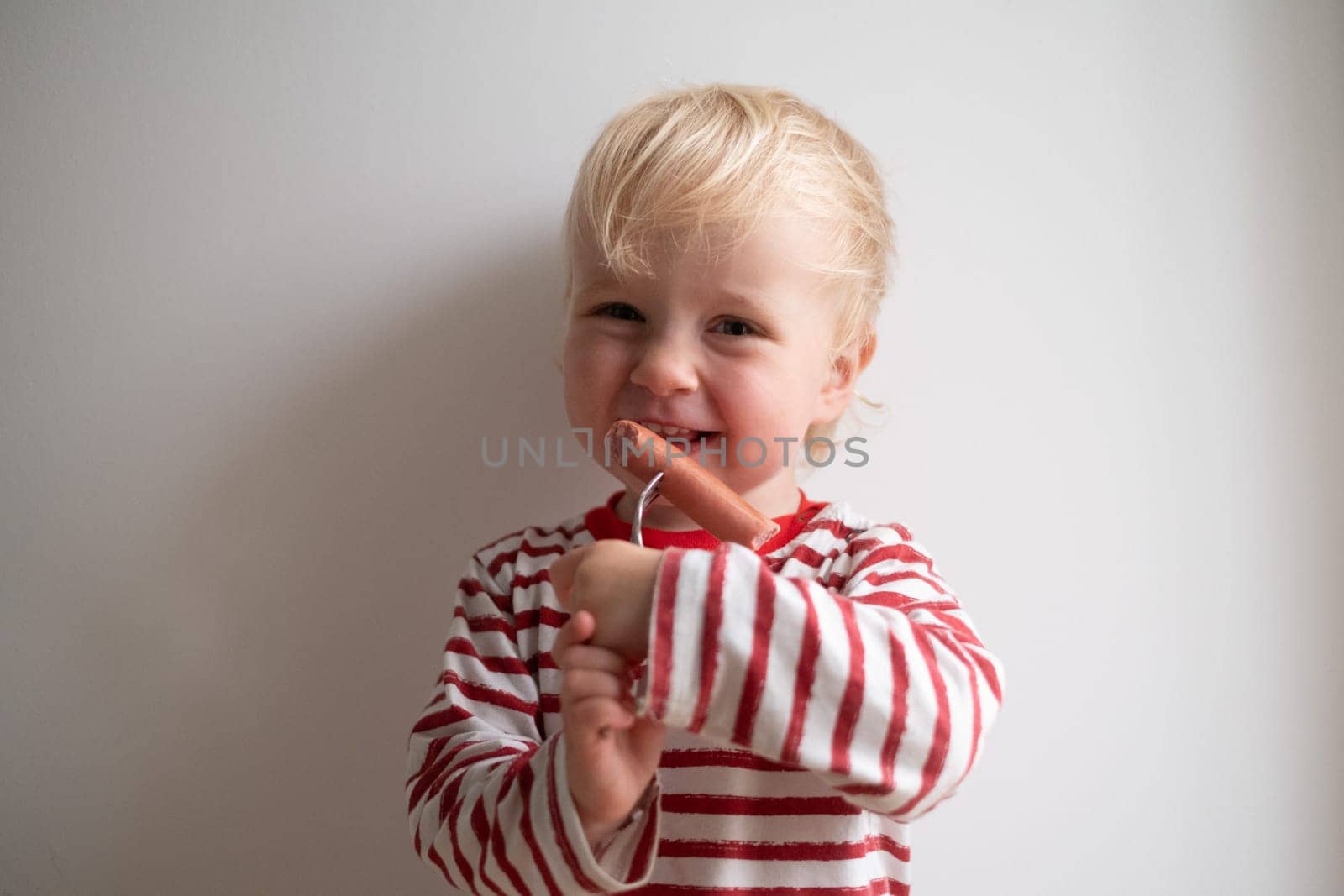 Little happy girl eating tasty sausage on white background.
