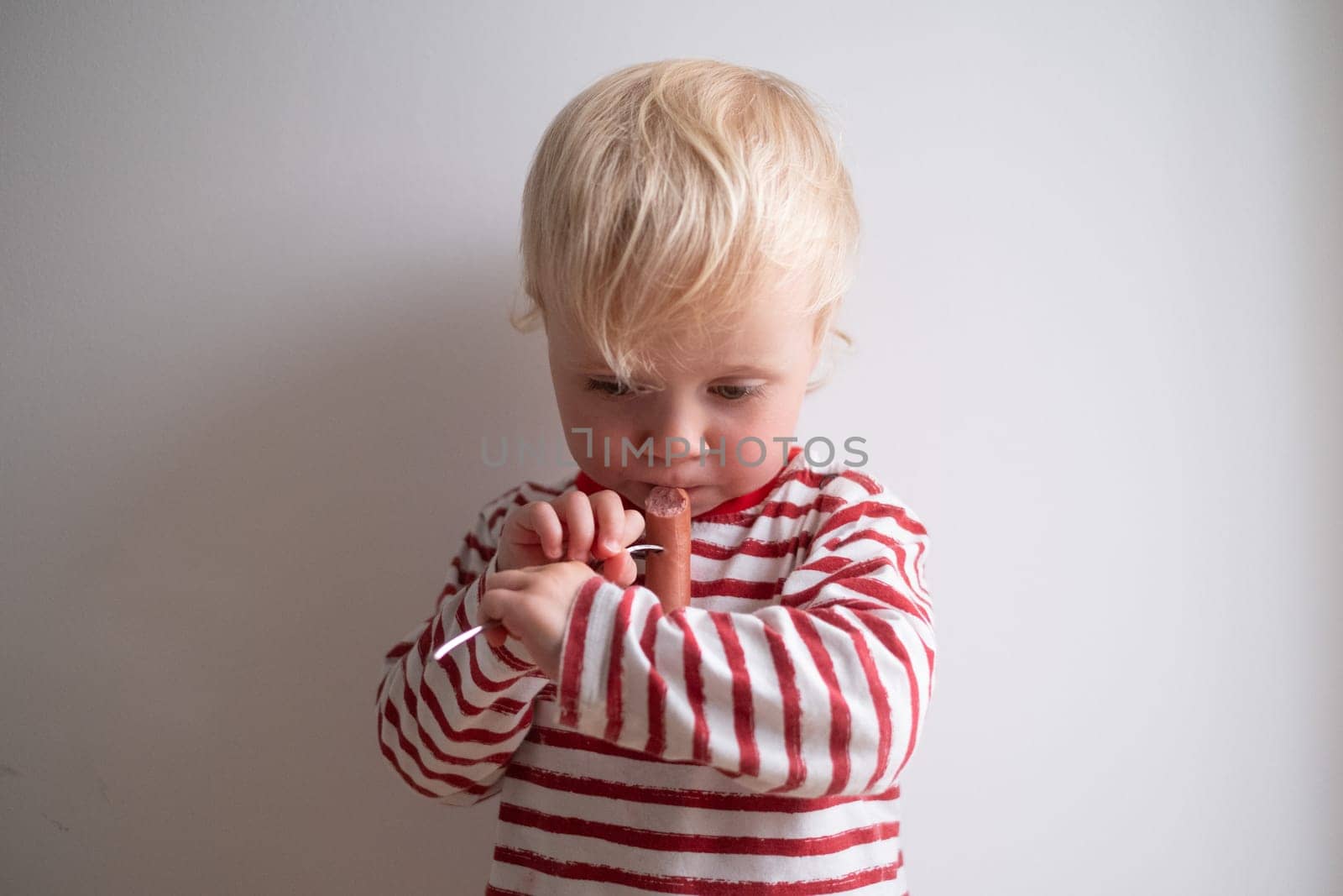 Little happy girl eating tasty sausage on white background.