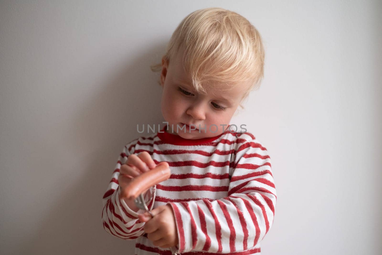 Little happy girl eating tasty sausage on white background.