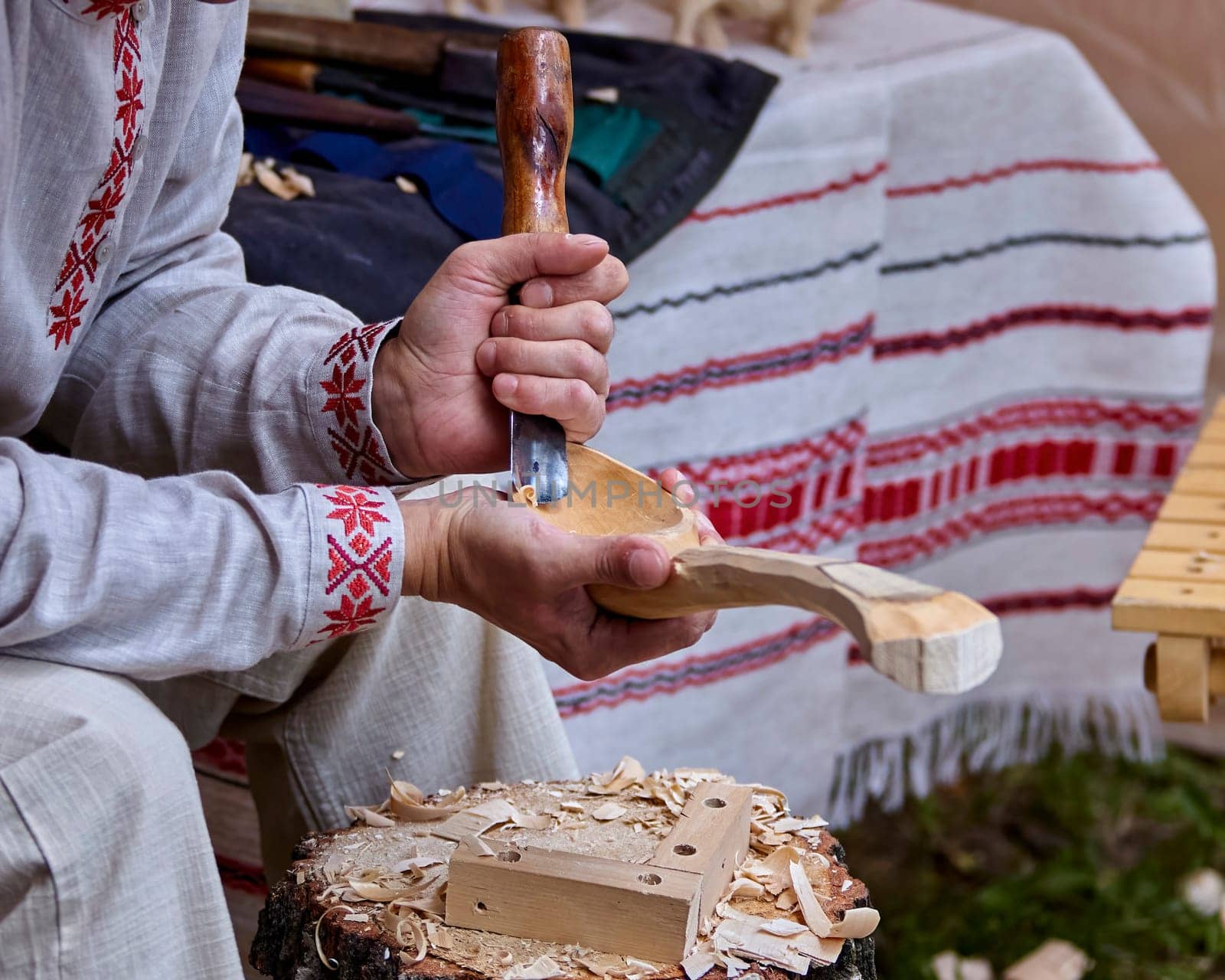 A craftsman, dressed in traditional attire, carefully carves a wooden spoon with a chisel and mallet. He is seated on a wooden stump, surrounded by the remnants of his work and the vibrant colors of a summer festival backdrop.