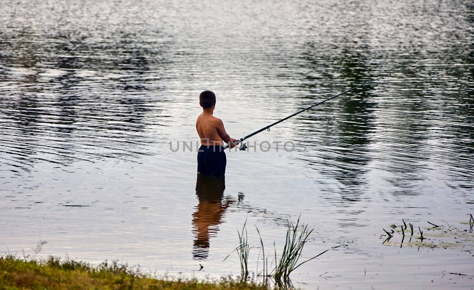 A young boy stands in a lake, holding a fishing rod and casting his line into the water. The sun reflects off the rippling surface.