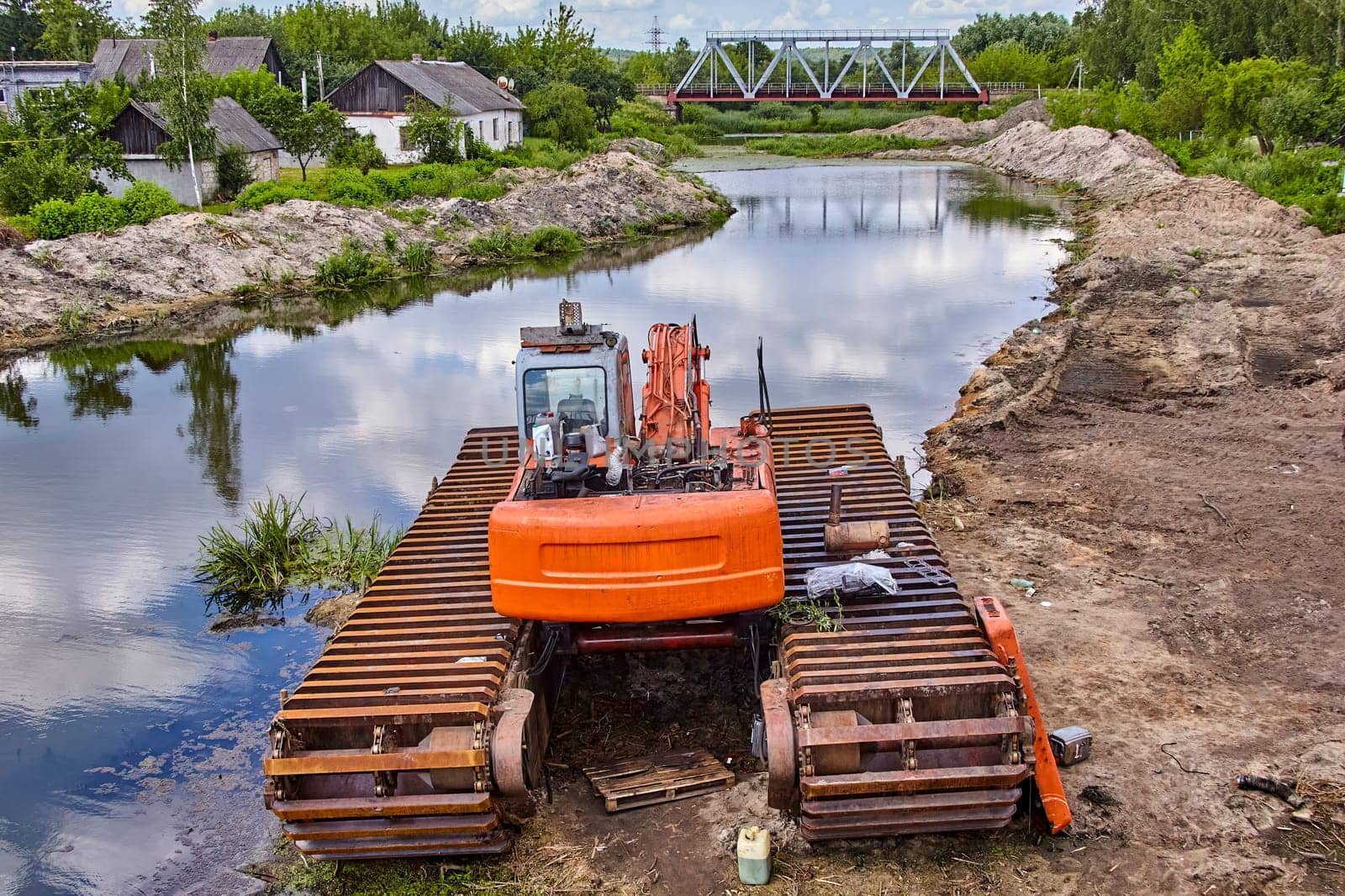 A bright orange amphibious excavator sits parked on the bank of a river, with a metal bridge in the distance.