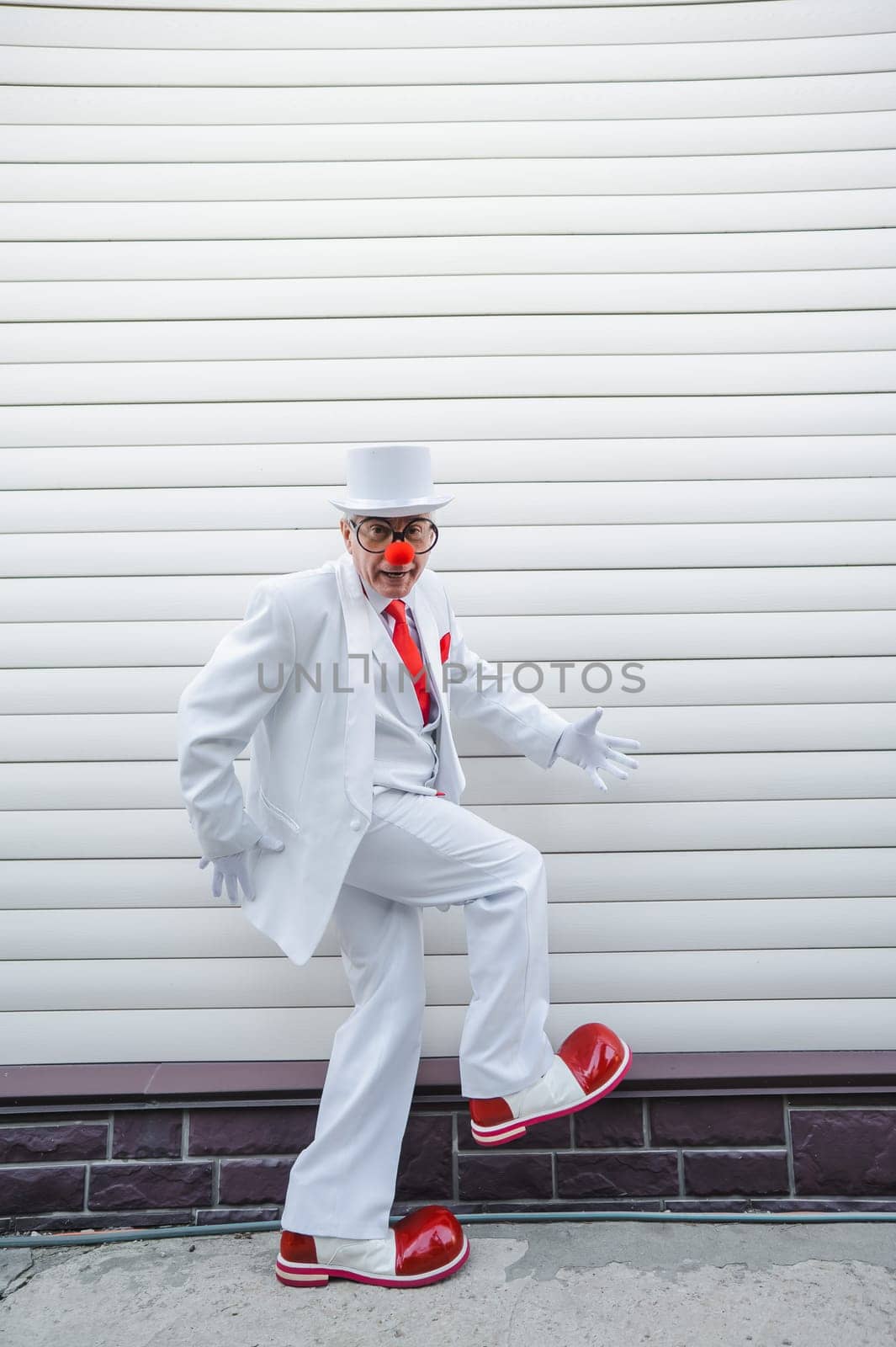 An elderly man in a white suit, huge boots and a clown nose walks funny