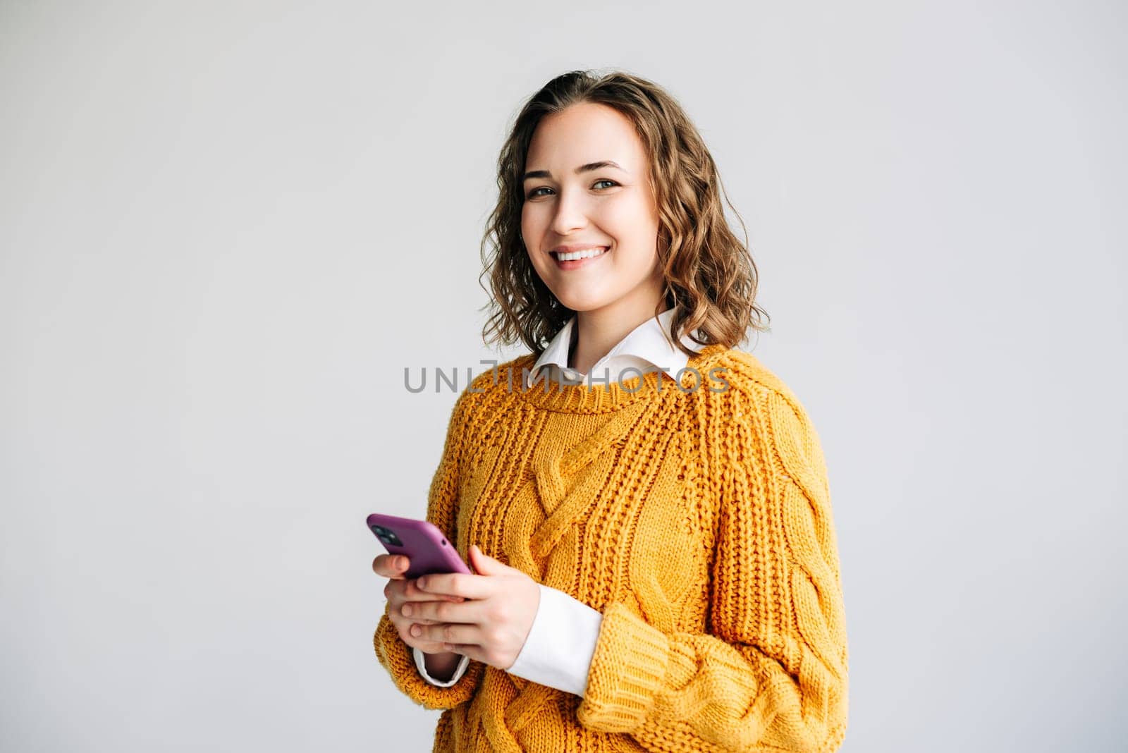 Smiling young woman with curly hair using her smartphone. She is browsing the internet, standing against a white background. Perfect for illustrating modern communication and technology usage.