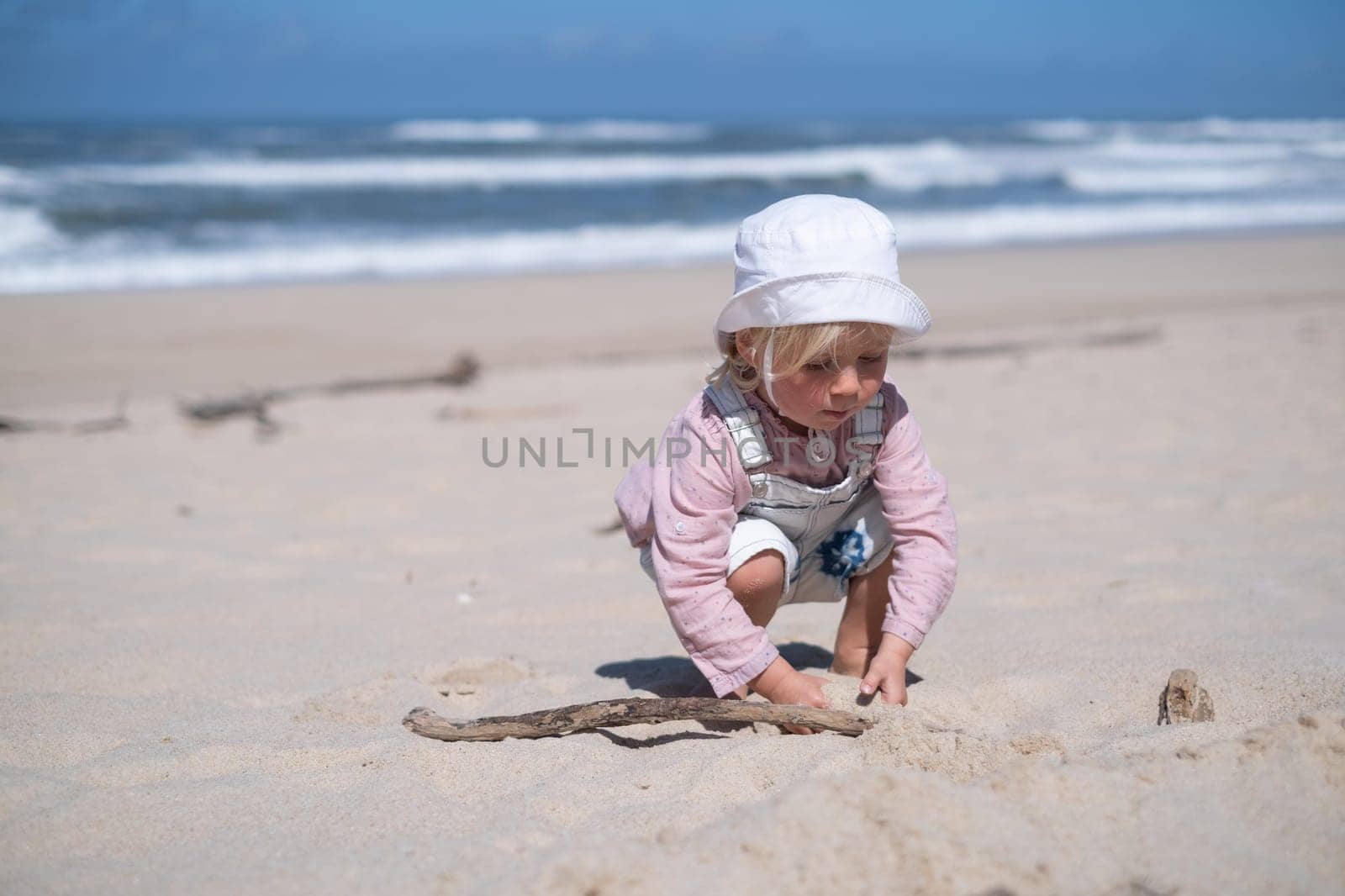Little girl is sitting on sand and playing at sea beach in warm sunny summer day