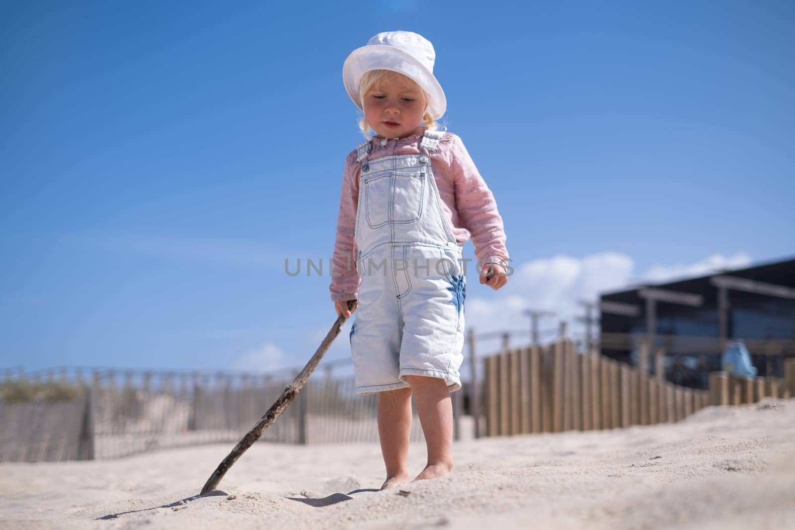 Little girl is sitting on sand and playing at sea beach in warm sunny summer day