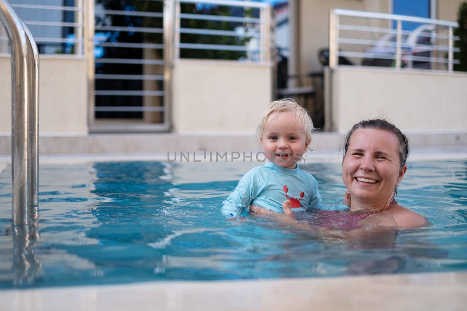 Mother and baby in outdoor swimming pool of tropical resort. Kid learning to swim. Mom and child playing in water. Family summer vacation in exotic destination. Active and healthy sport for kids.