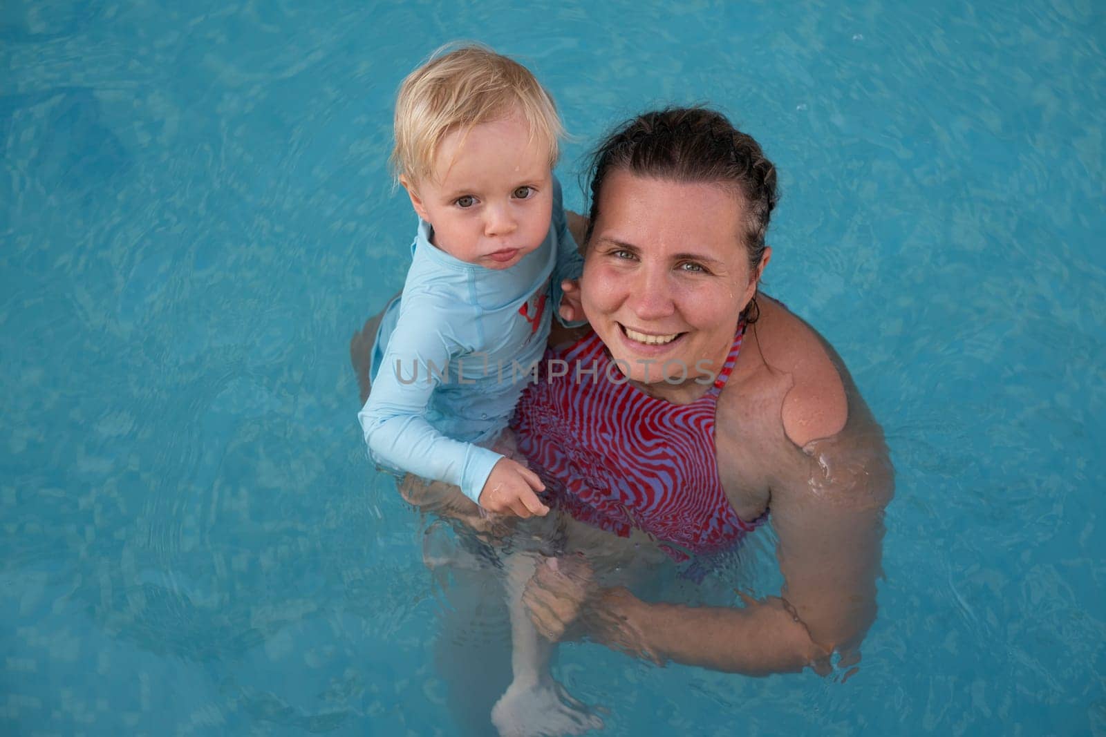 Mother and baby in outdoor swimming pool of tropical resort. Kid learning to swim. Mom and child playing in water. Family summer vacation in exotic destination. Active and healthy sport for kids.