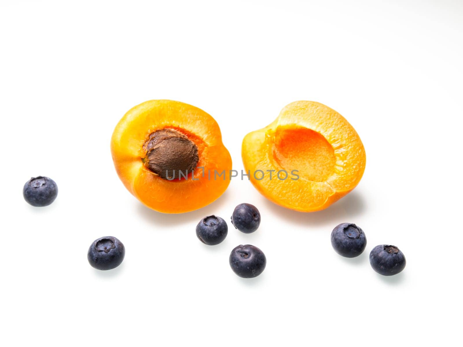 A close up food photo of an apricot cut in half displayed with the pit and some ripe blueberries.