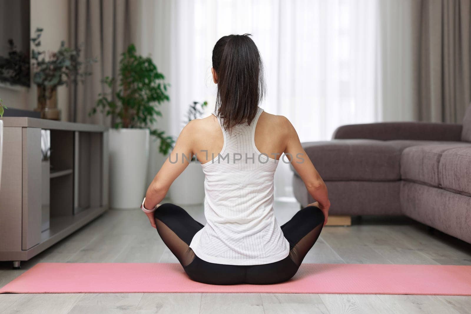 Back view of sporty woman practicing yoga and meditation sitting in lotus pose on pink yoga mat at home