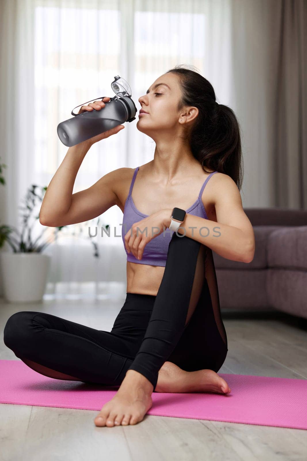 sporty woman drinking water from water bottle sitting on pink exercise mat at home