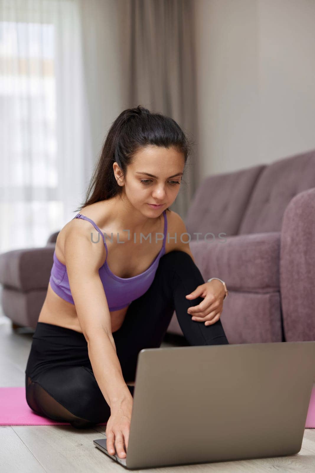 sporty Caucasian woman looking at laptop while sitting on pink fitness mat, ready for online yoga training at home.