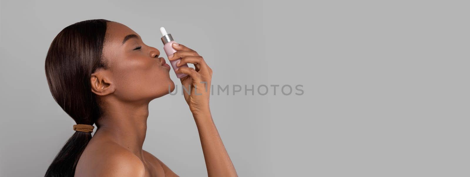 Happy pretty black woman with perfect pure skin kissing jar of oil, isolated on gray background, profile, copy space, studio. Anti-aging serum for treatment and natural cosmetics, body and beauty care