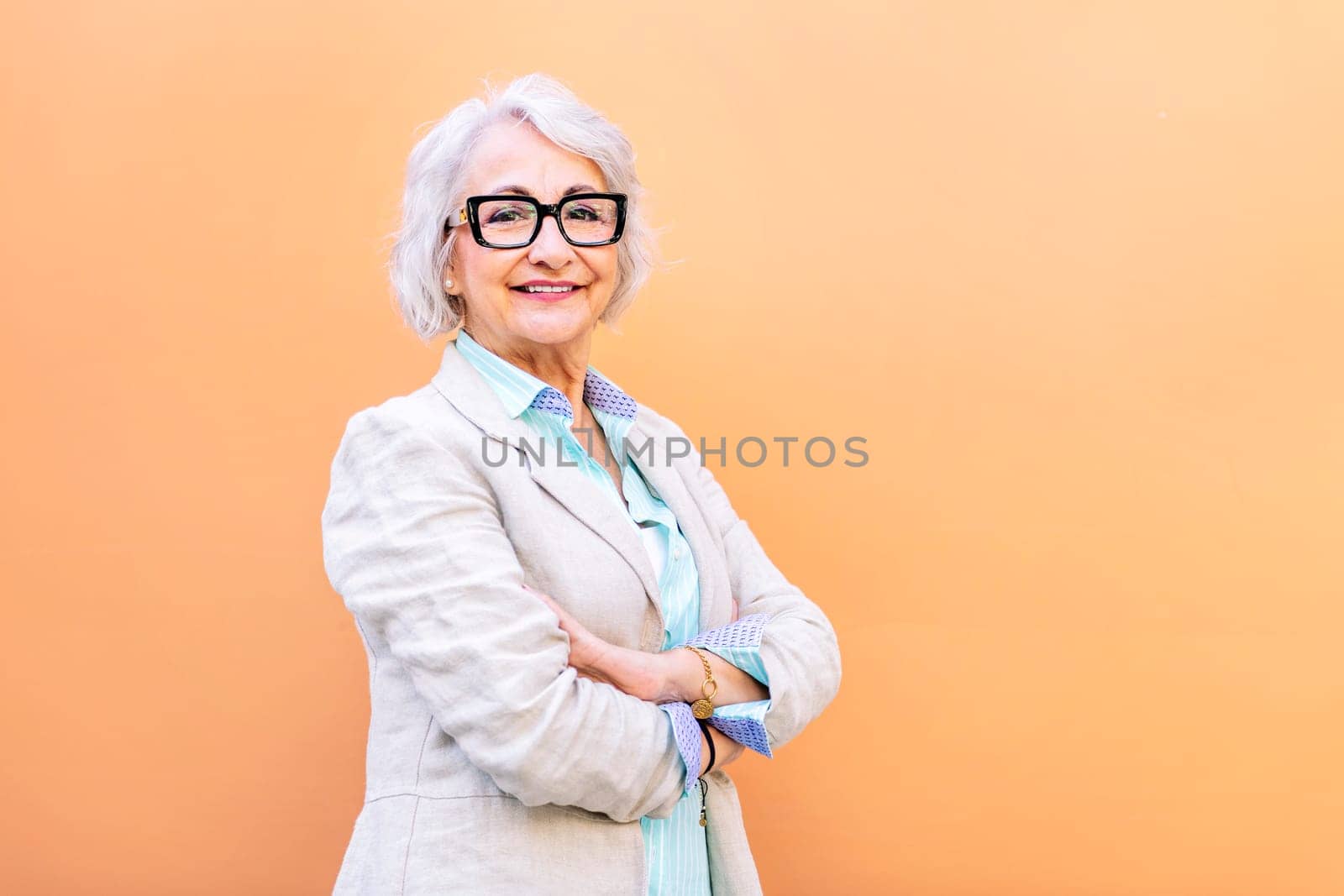 stylish mature entrepreneur woman smiling happy looking at camera leaning on an orange wall with arms crossed, concept of active lifestyle in adulthood, copyspace for text