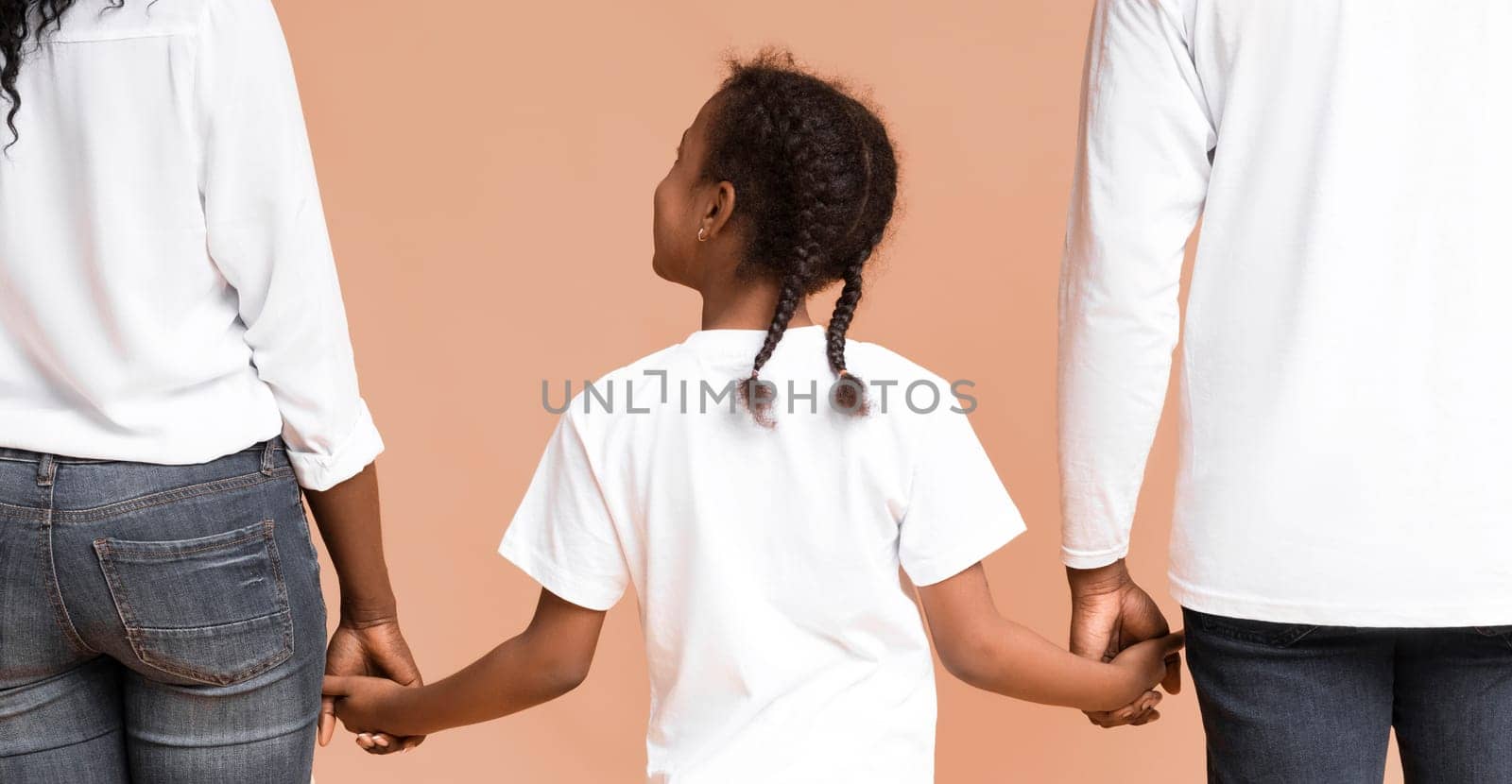 Family support. Little black girl with braids holding hands of her mom and dad while they walking together over yellow background, rear view