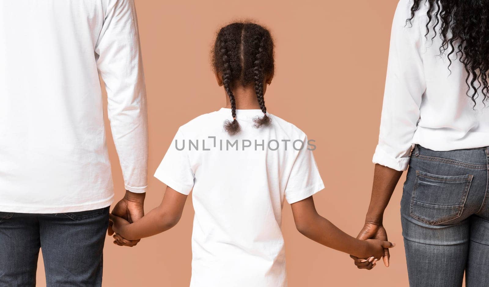 Adoption concept. Little afro girl holding hands of her adoptive parents, walking together over yellow background, rear view