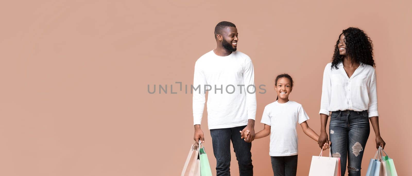 Family Shopping Concept. Happy African American parents and their daughter walking with shopper bags and holding hands in studio, yellow background.