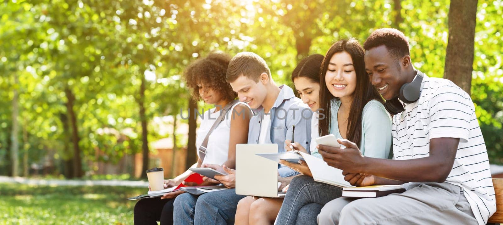 Education concept. Cheerful university students preparing for exam, talking and drinking coffee outdoors, sitting on bench in campus, free space