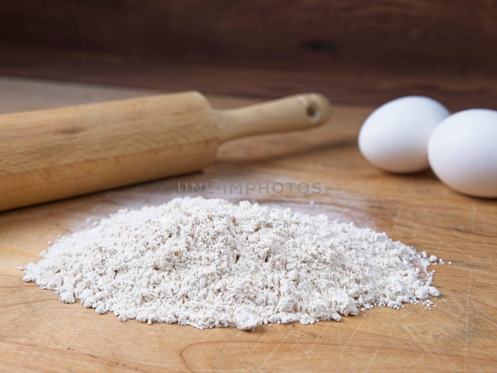 A close up of a pile of flour along with eggs and a rolling pin on a wooden cutting board.