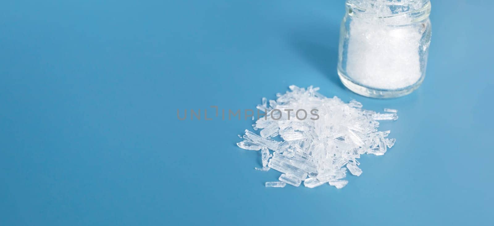 Banner Top View Organic Heap of Translucent Menthol Crystals on Blue Background, Glass Jar. Natural Ingredient for Producing Cosmetics. Horizontal Design Copy Space. High quality photo