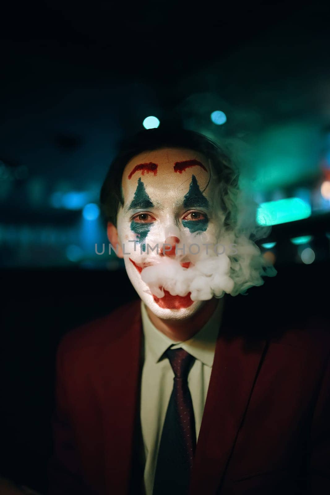 Person in suit and tie with the Joker makeup puffs smoke out of his mouth. Halloween party in nightclub with decorations and scary costumes. Autumn holiday. All Saints' Night.