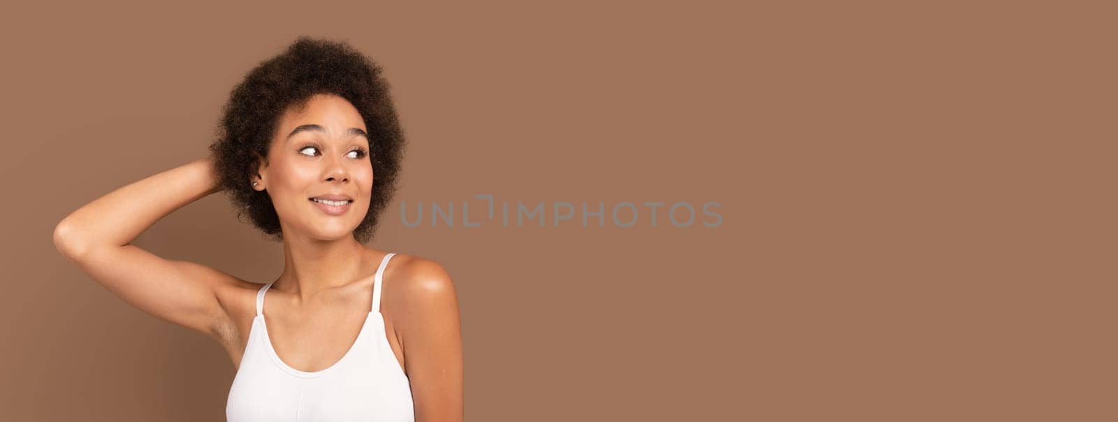 Cheerful millennial african american curly woman in white top with perfect skin looks at free space, isolated on brown background, panorama. Lady enjoy spare time, cosmetology, beauty care at home