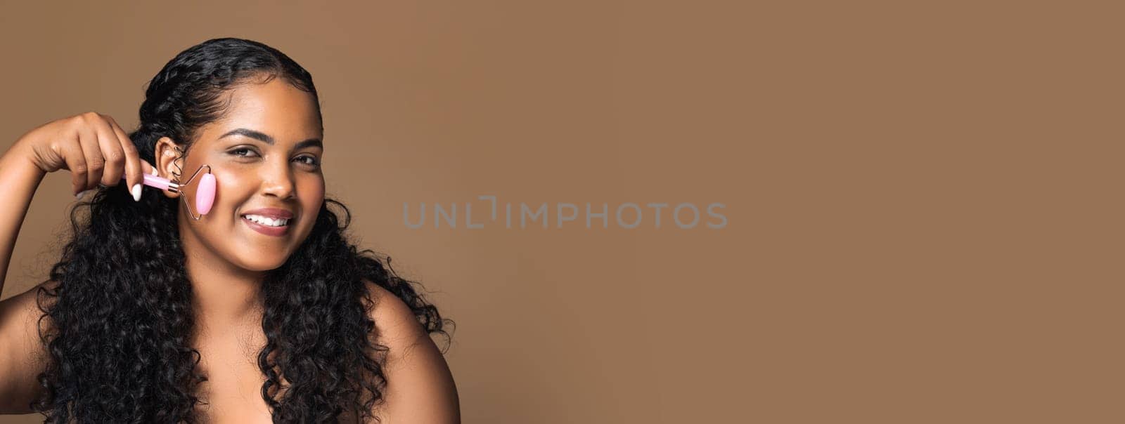 Happy brazilian oversize lady doing jade massage to face with quartz roller, smiling at camera, posing isolated on brown background, copy space. Beauty care, anti-wrinkle procedure