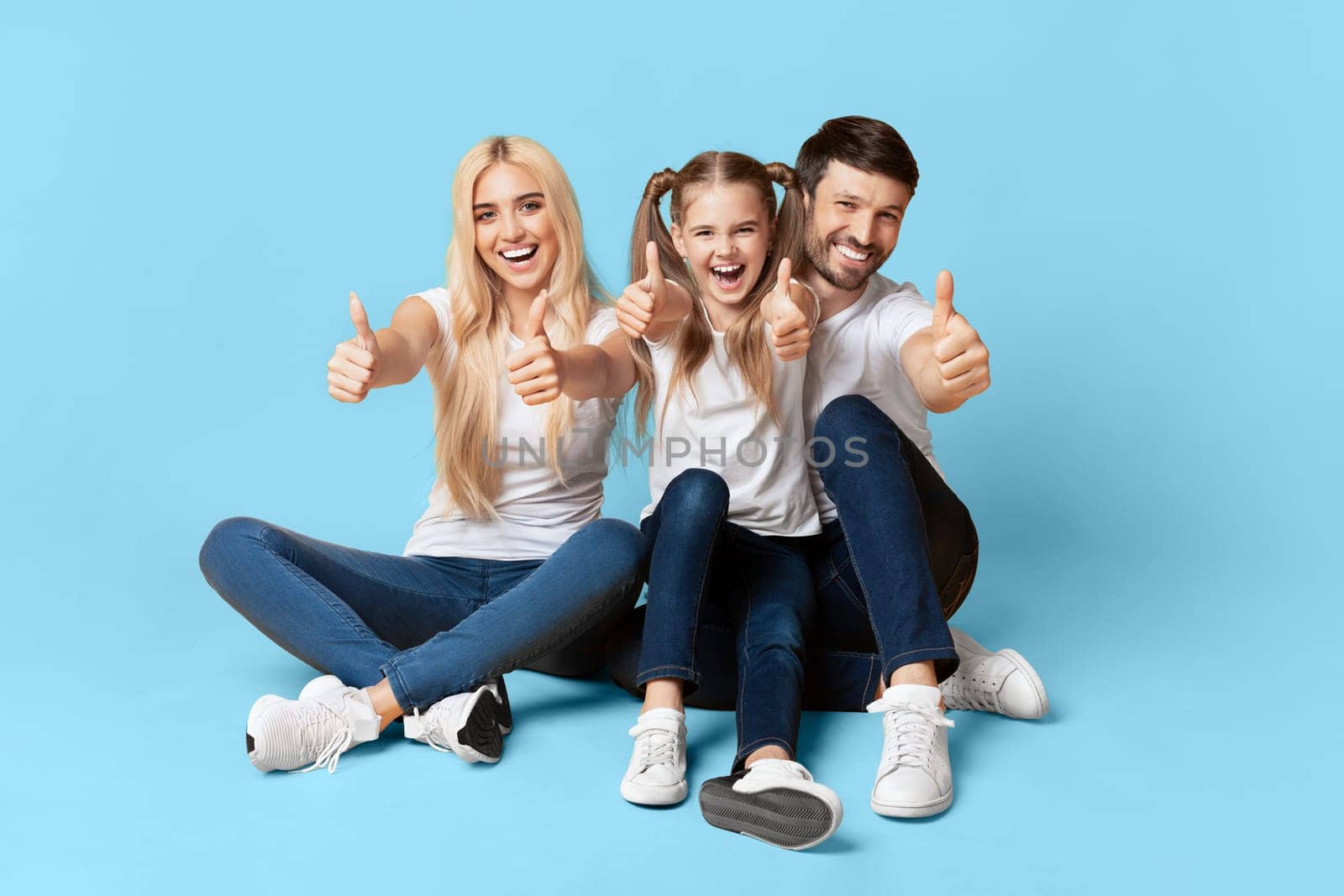 A family of three, a mother, father, and their young daughter, are sitting on a light blue background. They are all smiling and giving the thumbs up.