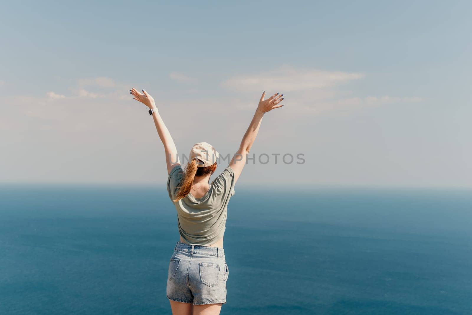 A young woman in a green shirt and denim shorts stands with her arms raised in celebration, enjoying the view of the blue ocean and the bright sky.