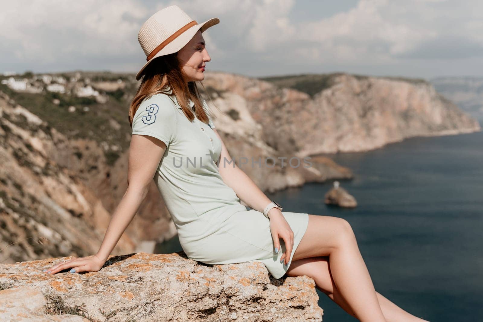 A woman wearing a light green dress and a straw hat sits on a rock cliff and looks out at the ocean.