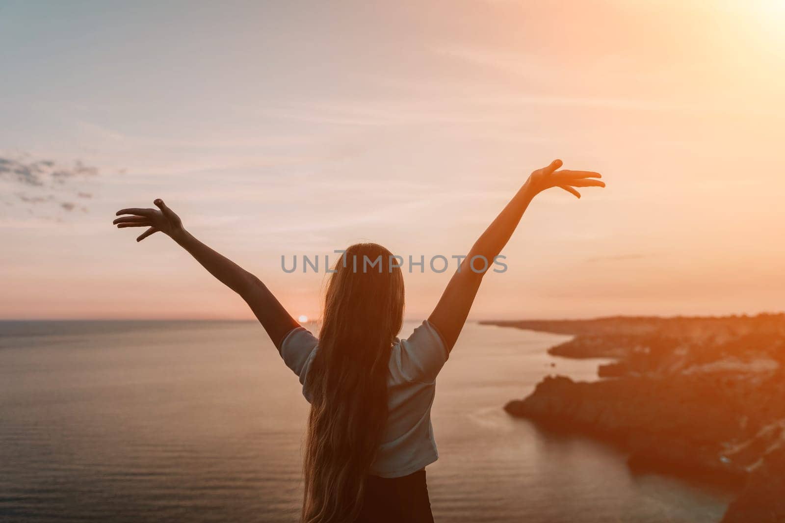 A woman stands with her arms raised in the air, silhouetted against a colorful sunset over the ocean.