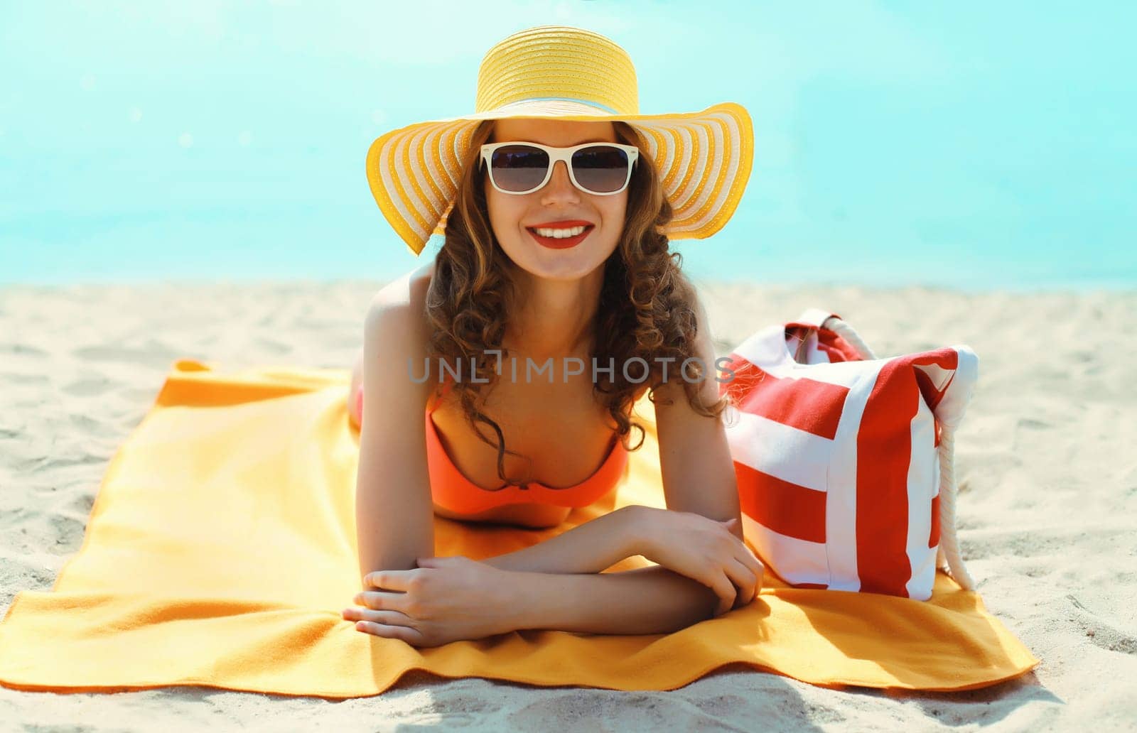 Summer vacation, happy smiling young woman with bag in bikini and straw hat lying on the sand on the beach on sea background