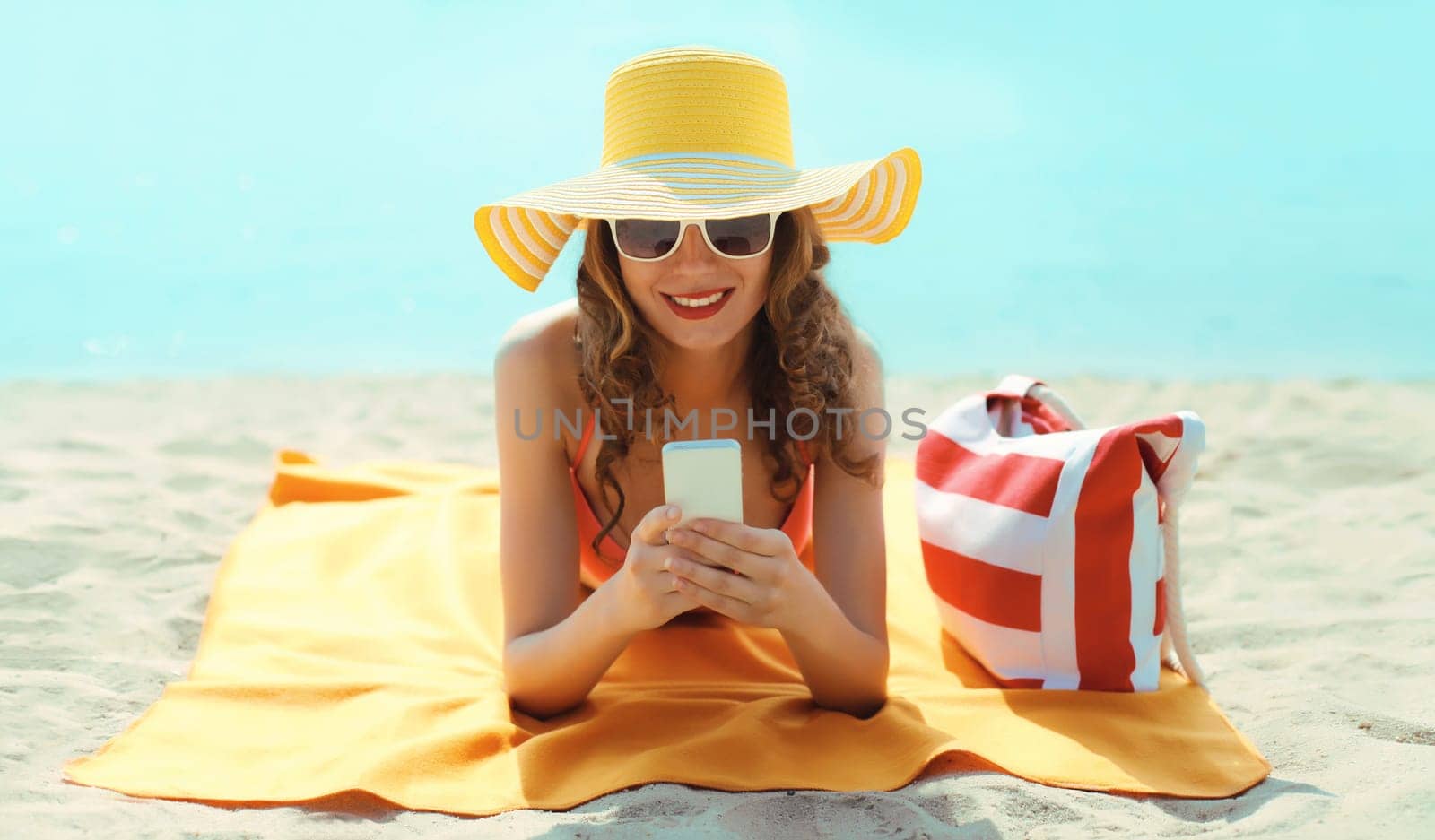 Summer vacation, happy young woman with phone, tourist hat, beautiful girl holding smartphone lying on the sand on the beach on sea background