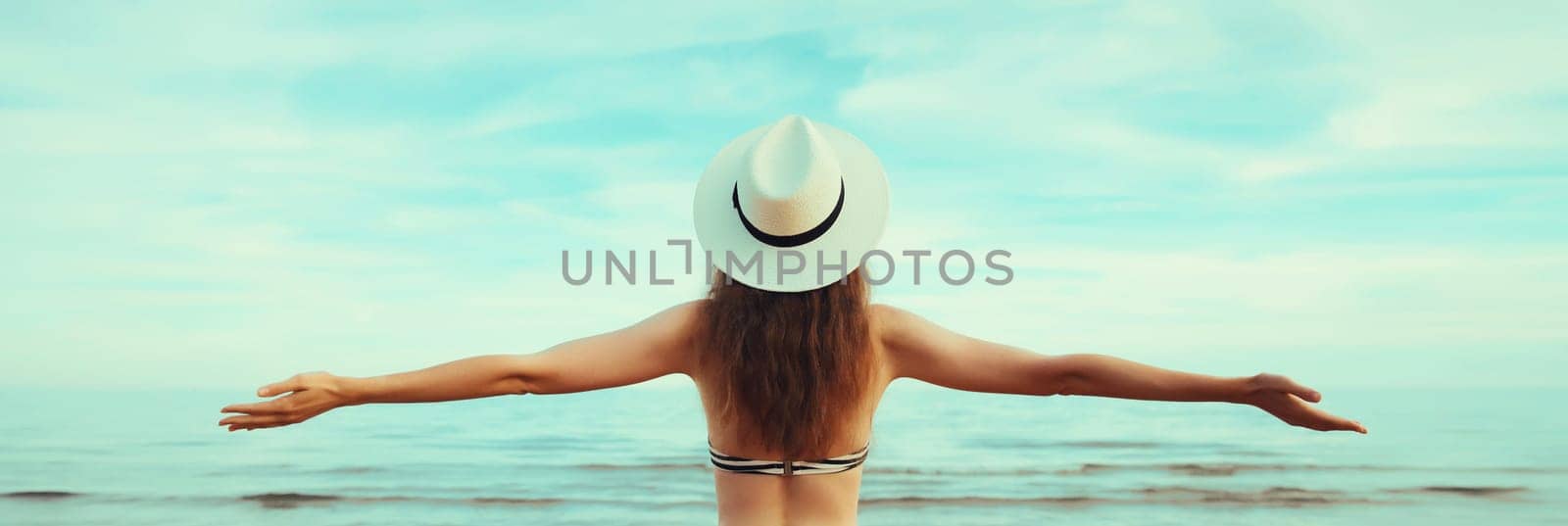 Summer vacation, rear view of beautiful happy woman in bikini swimsuit and tourist straw hat raising her hands up on the beach on sunny day on sea coast