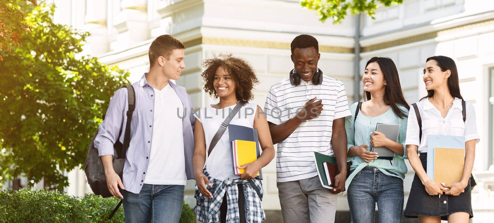 Group of multiethnic students walking together outdoors in college campus, holding books and notepads and laughing, copy space