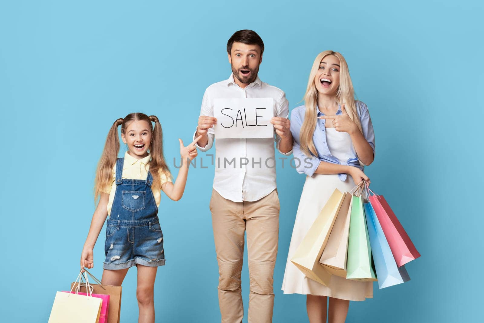 A family of three stands in front of a blue background, excitedly holding shopping bags and a sign that says Sale. The father holds the sign, the mother holds multiple shopping bags