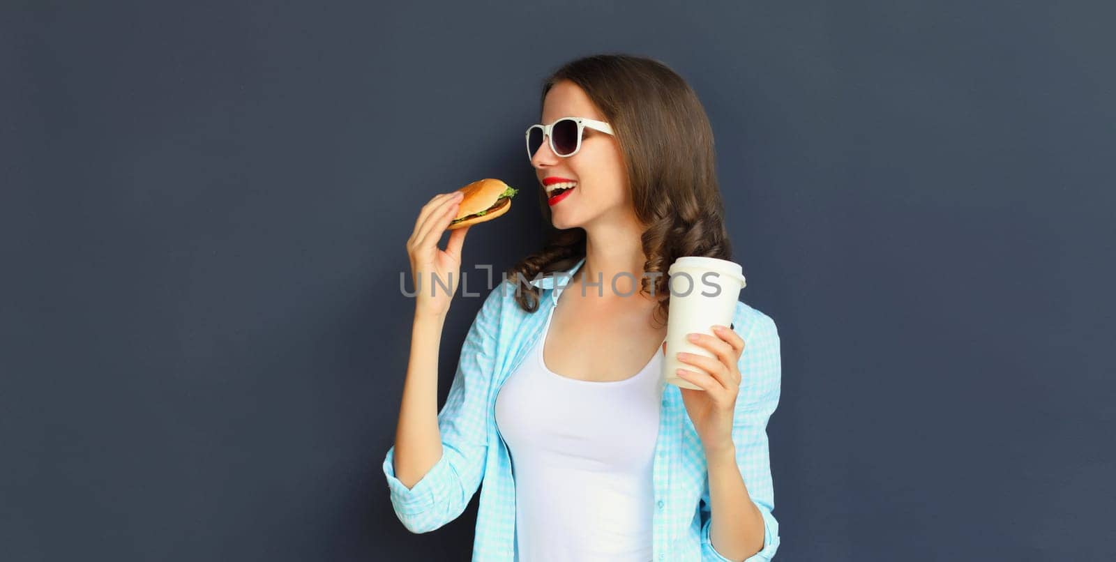 Portrait of happy cheerful young woman eating burger fast food holding cup of coffee, teenage girl having fun isolated on black wall studio background