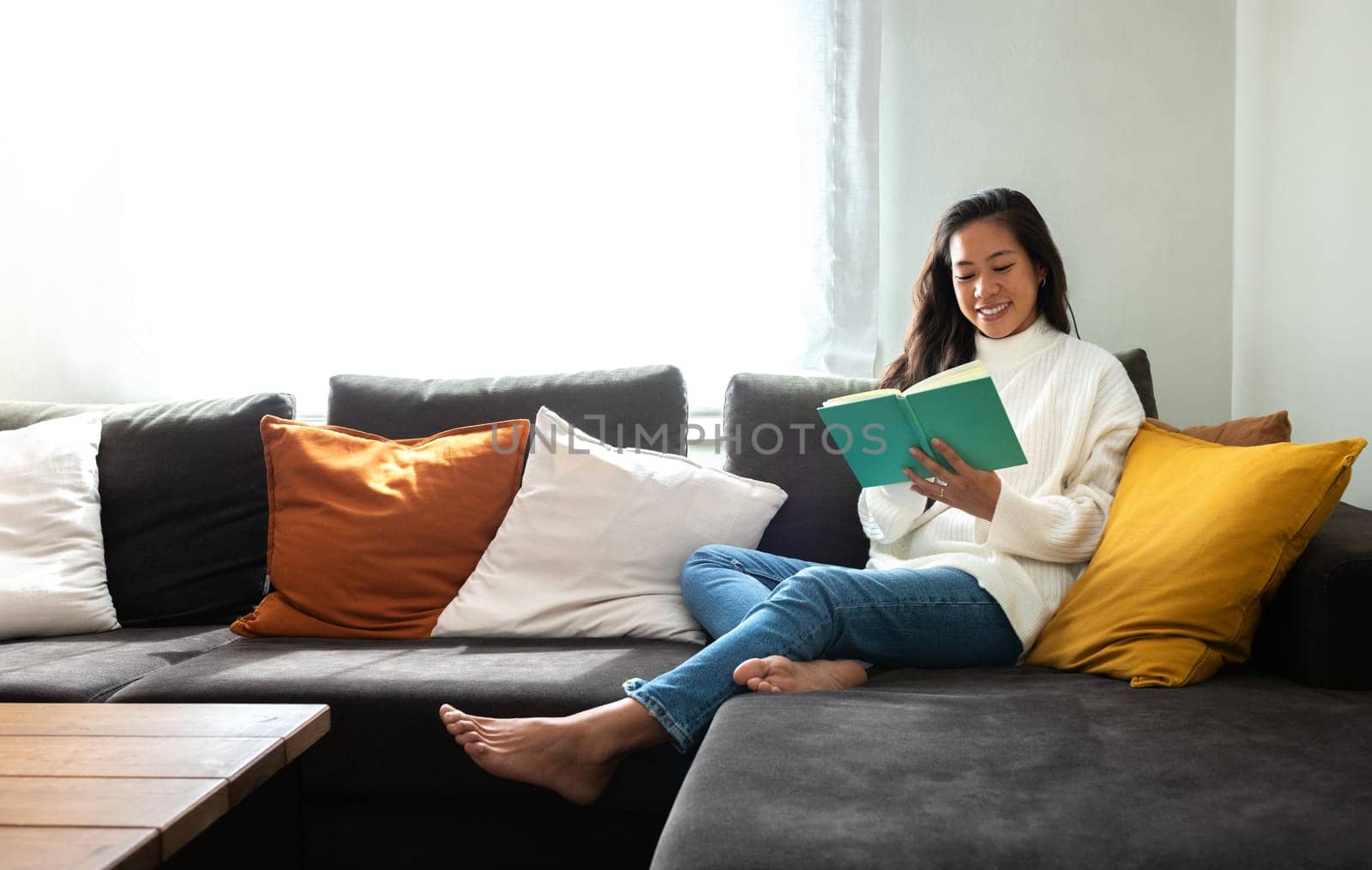 Happy, smiling Asian woman relaxing at home reading a book sitting on the sofa. Copy space. Lifestyle concept.