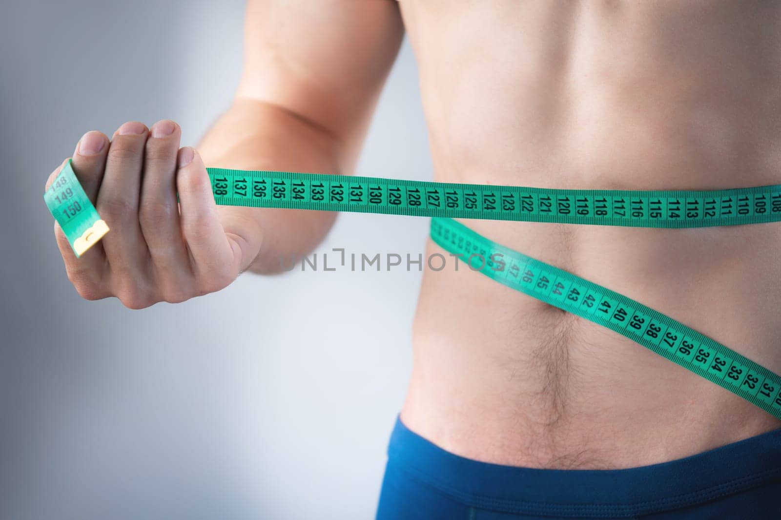 A sporty attractive man in underwear with a perfect body and a beautiful torso stands on a gray background, holding a tape measure on his thin waist to show the result of his workout. Weight loss concept.