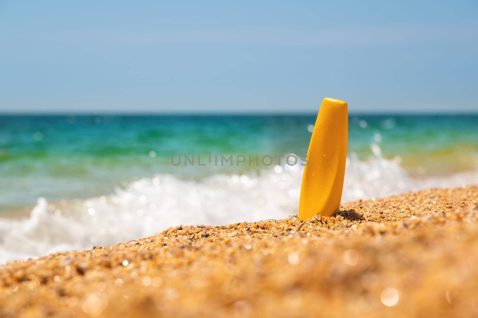 Yellow bottle of sunscreen on a golden beach next to the waves of the sea and foam against the skyline on a sunny day in summer. Mockup bottle without logo.