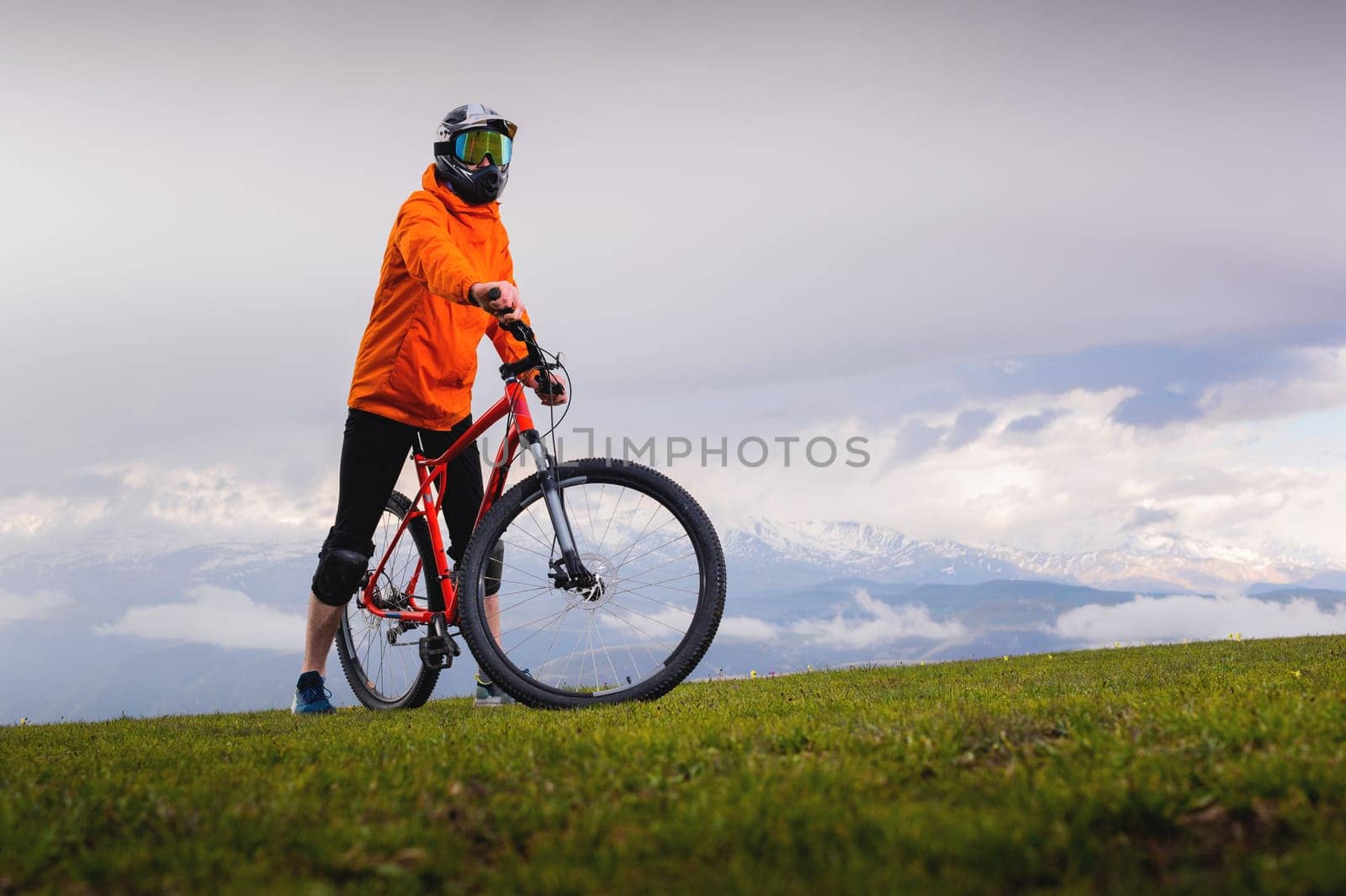 Portrait of a young cyclist standing against the backdrop of a summer landscape with a mountain in the clouds. A cyclist on a mountain path stops to watch the scenery of peaks in the distance by yanik88
