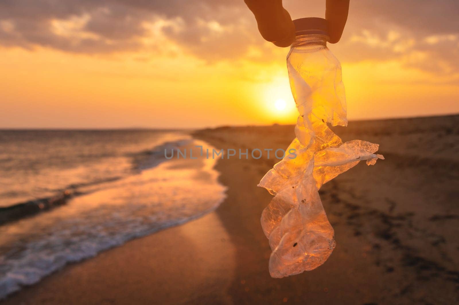 Man's hand picking up an empty plastic cleaning bottle on the beach, volunteer concept. Fingers hold an old bottle thrown out by the sea at sunset. Environmental pollution. Ecological problem by yanik88
