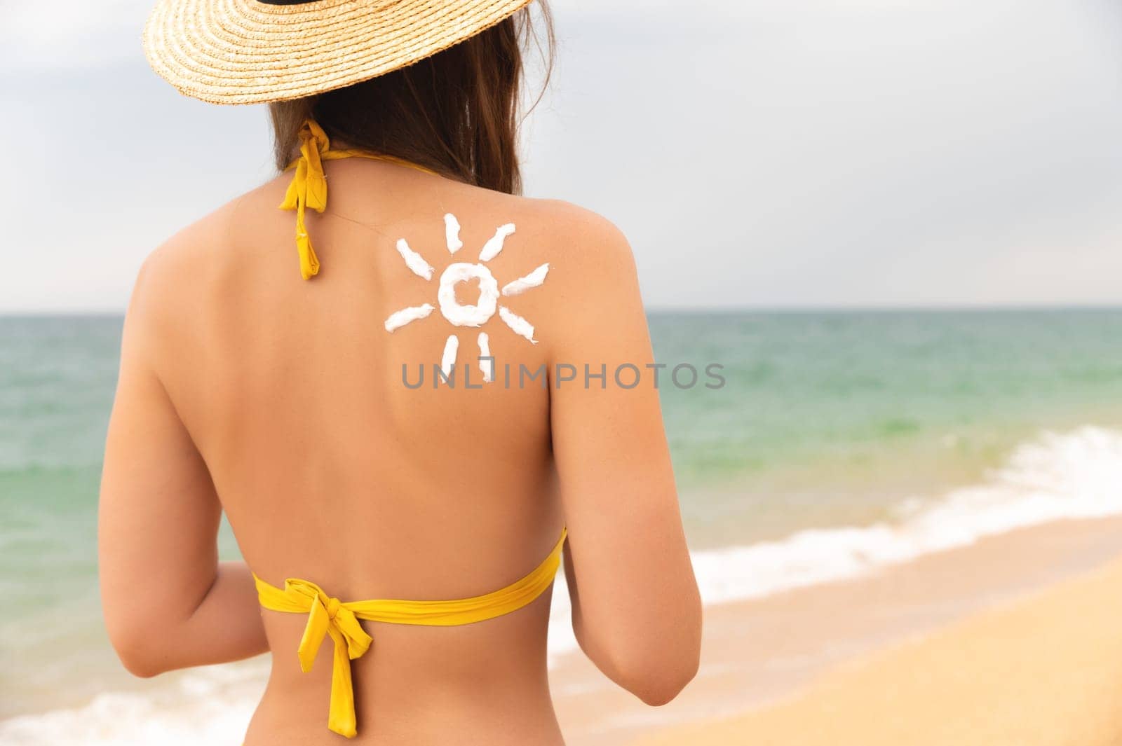 Sun protection. A beautiful, unrecognizable girl wears sunscreen on her tanned shoulder in the shape of a sun. Skin care, young woman in a straw hat and swimsuit stands with her back to the camera at sea.