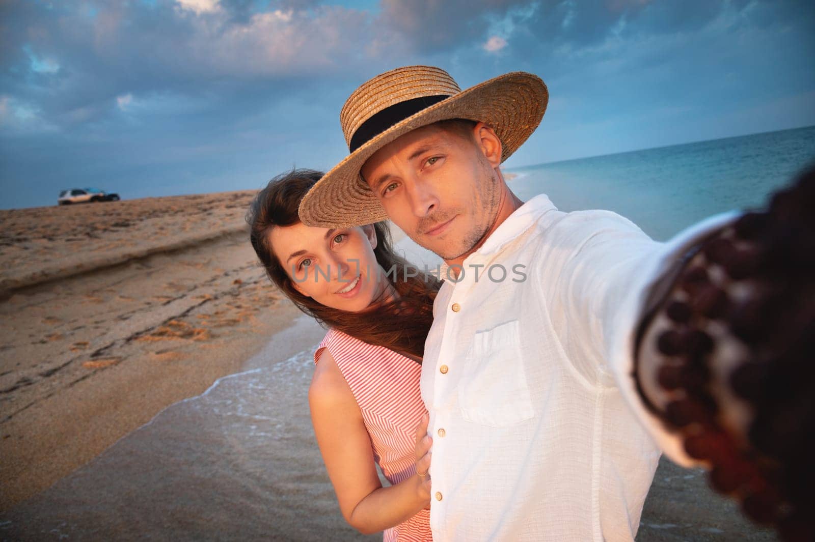Close-up of smiling happy young couple, two family man and woman hugging each other, taking first person selfie at sunrise or sunset on sea beach on summer day.
